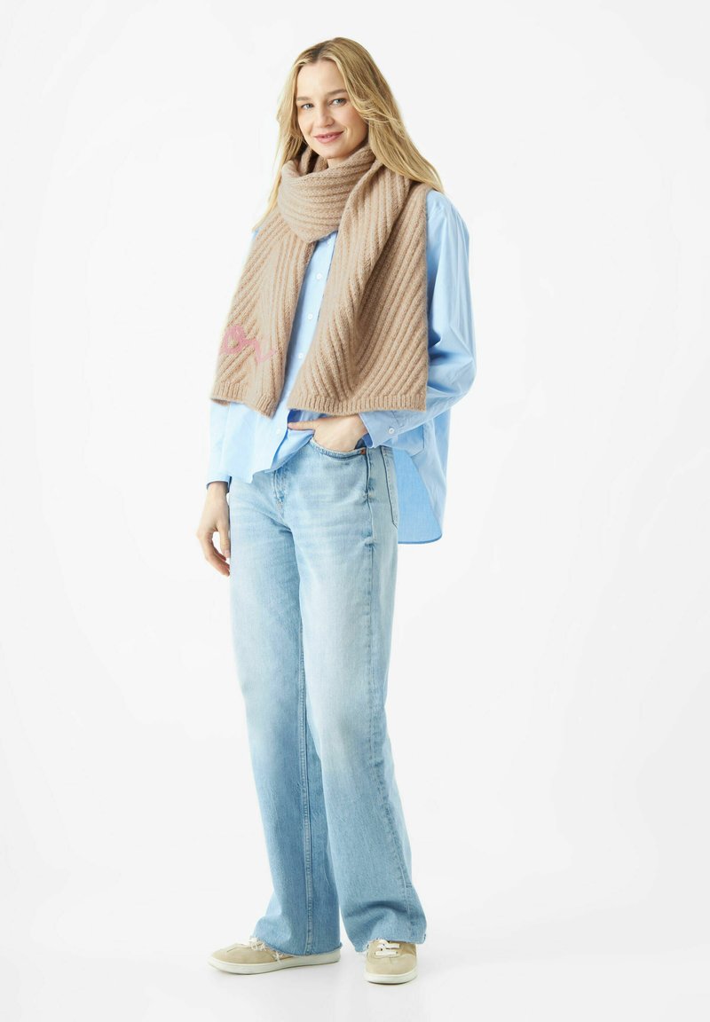 Beige gestrickter Schal mit diagonaler Rippenstruktur und rosa Stickerei. Getragen über einem hellblauen Hemd mit Knöpfen und lockeren blauen Jeans.