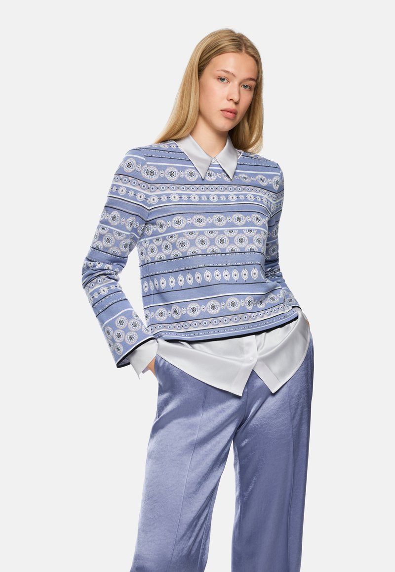 Light blue patterned sweater with a collared design, featuring intricate circular motifs and contrasting stripe details. Paired with satin trousers.