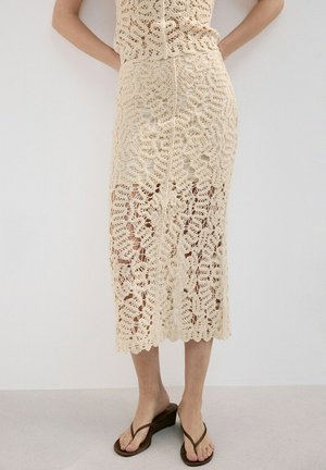 Jupe midi beige en crochet portée par une personne debout contre un mur clair uni, associée à des tongs compensées marron.