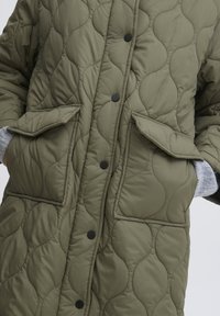Olivengrøn quilted jakke med et tekstureret mønster, featuring knaplukning foran, to frontlommer og sorte trykknapper.