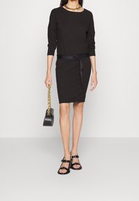 Schwarzes Baumwollkleid mit langen Ärmeln in taillierter Form, verstellbarem Gürtel und Logodetail. Kombiniert mit schwarzen Sandalen und einer schwarzen Handtasche mit Kettenriemen.