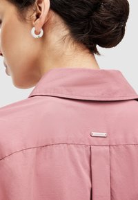 Camisa de color rosa con cuello y pliegue en la parte posterior, y etiqueta de logo. Presenta una textura suave y un brillo sutil con un diseño clásico de botones.
