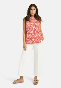 Rotes und weißes blumiges, kragenloses ärmelloses Oberteil kombiniert mit weißen weiten Hosen und beigen Riemchensandalen, was einen hellen, sommerlichen Look präsentiert.