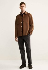 Giacca camicia di lana marrone con due tasche anteriori, chiusura con bottoni e una vestibilità leggermente over-size, abbinata a pantaloni neri e scarpe scure.