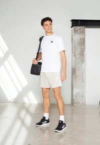 Λευκό κοντομάνικο μπλουζάκι με μαύρο λογότυπο, ανοιχτό μπεζ σορτς με πλαϊνές ρίγες, μαύρα αθλητικά παπούτσια και κρατώντας μια μαύρη τσάντα ώμου.