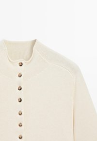 Krémově zbarvený pletený kardigan s kulatými hnědými knoflíky po celé přední části a mírně zvýšeným límcem.