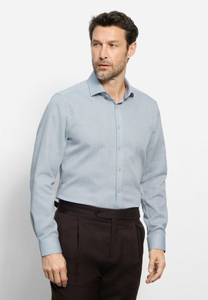 Homme portant une chemise bleu clair à manches longues boutonnée et un pantalon marron foncé, regardant vers sa gauche sur un fond clair uni.