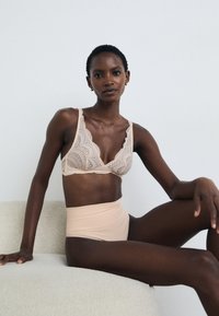 Bralette bege em renda com copas em forma de V e bordas onduladas, combinado com calcinhas bege de cintura alta. Textura suave e padrão delicado.