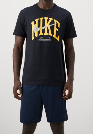 Homme portant un t-shirt noir Nike avec un logo jaune et un short bleu marine, debout devant un fond uni.