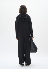 Felpa nera oversize con cappuccio, abbinata a pantaloni larghi neri e una borsa a spalla nera, caratterizzata da una texture liscia e un design minimale.
