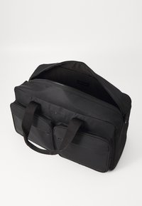 Svart stoffpose med to ytre glidelåslommer, dobbelhåndtak og glidelås lukking, med en glatt tekstur og minimalistisk design.