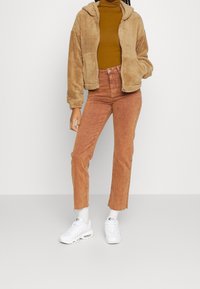 Giacca in pile marrone con zip, dolcevita senape, jeans a gamba dritta color ruggine e sneakers bianche con dettagli neri.