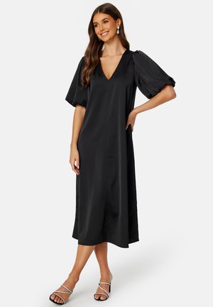 Abito midi in satin nero con scollatura a V, maniche a puff corte e una texture liscia. Abbinato a tacchi neri con cinturini.