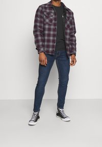 Camisa de cuadro en púrpura oscuro y gris, camiseta negra debajo, vaqueros ajustados azules y zapatillas negras de caña alta sobre un fondo liso.