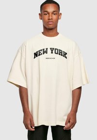 Creme oversized T-shirt med "NEW YORK" i fed sort tekst og "MERCHCODE" logo nedenunder, med korte ærmer og en afslappet pasform.