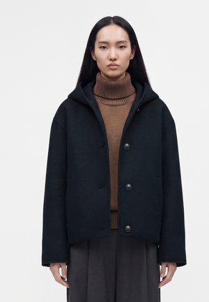 Jeune femme aux longs cheveux noirs portant un pull à col roulé marron et un manteau noir à capuche avec trois boutons, debout face à l’avant.