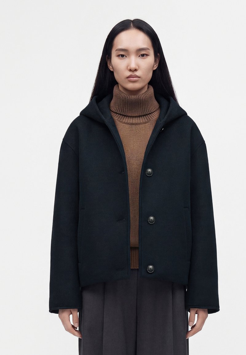 Jeune femme aux longs cheveux noirs portant un pull à col roulé marron et un manteau noir à capuche avec trois boutons, debout face à l’avant.