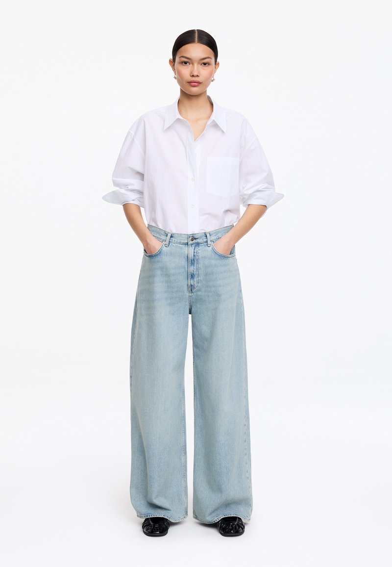 Femme aux cheveux foncés portant une chemise blanche boutonnée, un jean large clair et des chaussures noires, debout les mains dans les poches devant un fond blanc.
