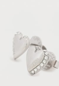 Ασημένια σκουλαρίκια σε σχήμα καρδιάς με στρας και χαραγμένο το λογότυπο "GUESS". Λείο πέτσωμα και γυαλιστερό φινίρισμα.