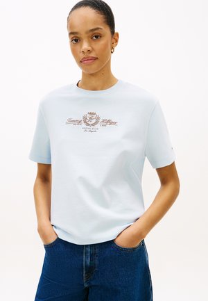 Kvinde med trukket hår bærer lyseblå Tommy Hilfiger t-shirt med logo og mørkeblå jeans, hænderne i lommerne, neutral baggrund.