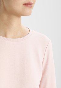 Felpa rosa chiaro con scollatura a costine, texture morbida e maniche lunghe, vista dal alto del corpo.