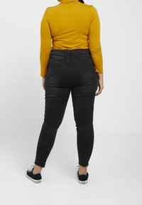 Vaqueros negros ajustados de talle alto, con dos bolsillos traseros y dobladillo deshilachado; combinados con un jerséi ajustado de cuello alto color mostaza y zapatillas negras.