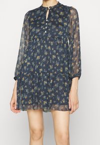 Navy chiffon-kukkakuosi mekko, jossa on rypytetty pääntie, avonainen kaula-aukko ja pitkät hihat. Kerroksellinen muotoilu, jossa on läpinäkyvä rakenne.