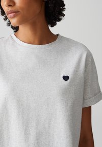 Graues Baumwoll-T-Shirt mit gerollten Ärmeln, ausgestattet mit einer kleinen, navyblauen Herzstickerei auf der linken Brustseite. Weicher Stoff, lockere Passform.