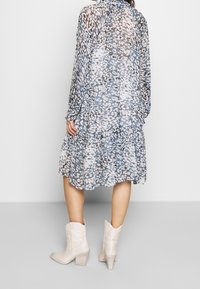 Plynulá dlouhá šaty s dlouhými rukávy s modro-bežovým abstraktním vzorem a vrstveným lemem. Kombinováno se světlými koženými cowboyskými botami na podpatku.