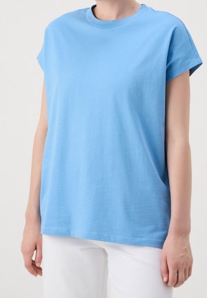 Person trägt ein locker sitzendes, himmelblaues T-Shirt mit kurzen Ärmeln und weiße Hose, steht vor einem schlichten hellen Hintergrund.