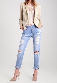 Blazer beige su una camicia button-up a righe multicolore, abbinato a jeans chiari strappati con risvolti.