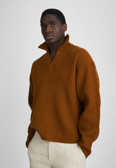 Pull côtelé en orange brûlé avec un col en V et un col surélevé ; coupe décontractée et manches à revers, associé à un pantalon beige clair.
