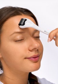 Derma roller med vitt handtag och svart microneedle-huvud, placerad på en panna mot en enfärgad vit bakgrund.