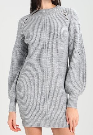 Donna che indossa un vestito maglione grigio con maniche a palloncino e texture a coste, in piedi davanti a uno sfondo chiaro e uniforme.