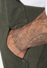 Olivgrüne Latzhose mit einer Tasche, geripptem Bund und sichtbaren Nähten. Nahaufnahme einer Hand, die in der Seitentasche ruht, um die Textur zu zeigen.