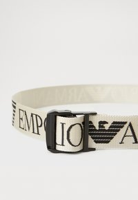 Beige geweven riem met zwart "EMPORIO ARMANI"-tekst en embleem. Het heeft een metalen gesp voor aanpassing en een gladde textuur.