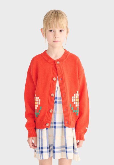 Niña joven con un cárdigan naranja abotonado con diseños de flores pixeladas sobre un vestido de cuadros azules y blancos.