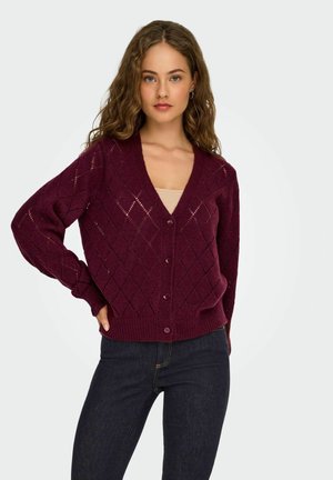 Bordeaux gebreide cardigan met een V-hals, diamantpatroon, knoopsluiting en lange mouwen. Gecombineerd met een beige tanktop en een donkere spijkerbroek.