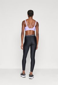 Fioletowy stanik sportowy z regulowanymi ramiączkami, w zestawieniu z czarnymi legginsami z wysokim stanem, która mają białą, markową tasiemkę w pasie oraz elegancki krój.