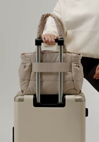 Sac fourre-tout beige rembourré maintenu sur une valise à roulettes argentée, doté de deux courtes poignées et d'une sangle centrale pour plus de stabilité.