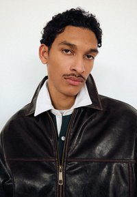 Young man with short curly hair and thin mustache wearing a white-collared shirt under a dark brown leather jacket against a plain background.