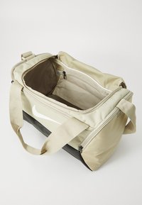 Sac de sport de taille moyenne, en tissu beige, avec un fond noir, arborant un grand logo Nike blanc, une bandoulière réglable et un compartiment extérieur zippé.
