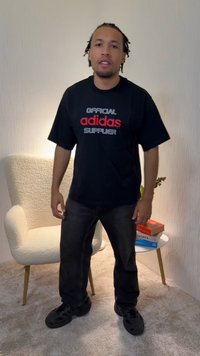 Juoda medvilninė marškinėliai su raudonu ir baltu užrašu „OFFICIAL adidas SUPPLIER“, derinami su tamsiomis džinsinėmis kelnėmis ir juoda avalyne, stovint jaukioje aplinkoje.
