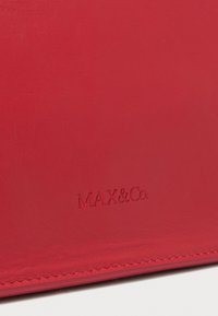 Rotes Lederprodukt mit sichtbarer Naht entlang der unteren Kante und geprägtem "MAX&Co."-Logo, das zentriert über der Naht platziert ist.