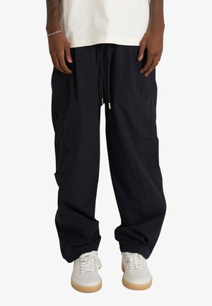 Persona che indossa pantaloni neri larghi con coulisse, sneakers bianche e una camicia bianca, in piedi davanti a uno sfondo semplice.