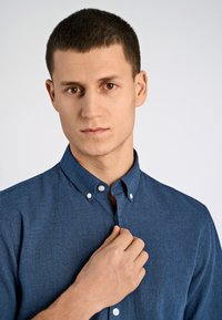 Koszula zapinana na guziki w ciemnoniebieskim materiale, z kołnierzykiem i białymi guzikami, o fakturowanym wykończeniu. Model poprawia kołnierzyk jedną ręką.