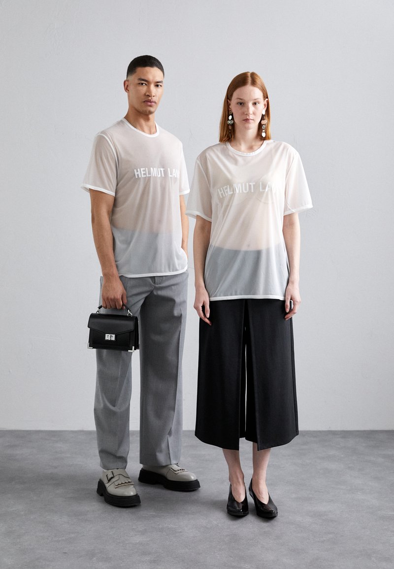 Transparente Mesh-T-Shirts mit dem Logo "HELMUT LANG", grauer Verlauf am unteren Rand. Das Model kombiniert helle und dunkle weit geschnittene Hosen mit kontrastierenden Schuhen.