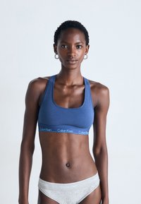 Kvinnelig modell iført blå Calvin Klein sports-BH og lys grå Calvin Klein undertøy, som står mot en enkel lys bakgrunn.