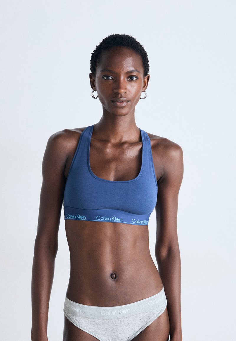 Kvinnelig modell iført blå Calvin Klein sports-BH og lys grå Calvin Klein undertøy, som står mot en enkel lys bakgrunn.