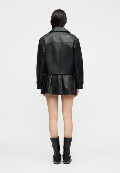 Γυναίκα με μακριά σκούρα μαλλιά, ντυμένη με μαύρο δερμάτινο μπουφάν, μαύρη πλισέ δερμάτινη μίνι φούστα και μαύρα μπότες μέχρι τη μέση της γάμπας με αλυσίδες, όρθια.